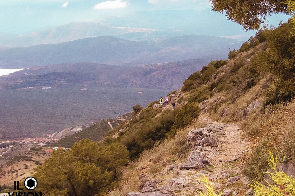 A mountainous landscape with a rocky path, dense vegetation, and distant hills.
