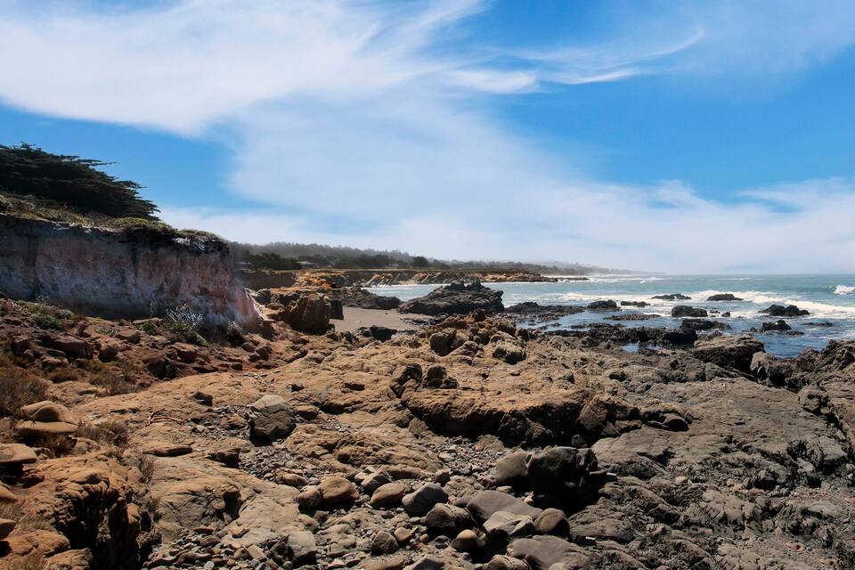 Um litoral rochoso com ondas quebrando na praia e um penhasco com vegetação rala.