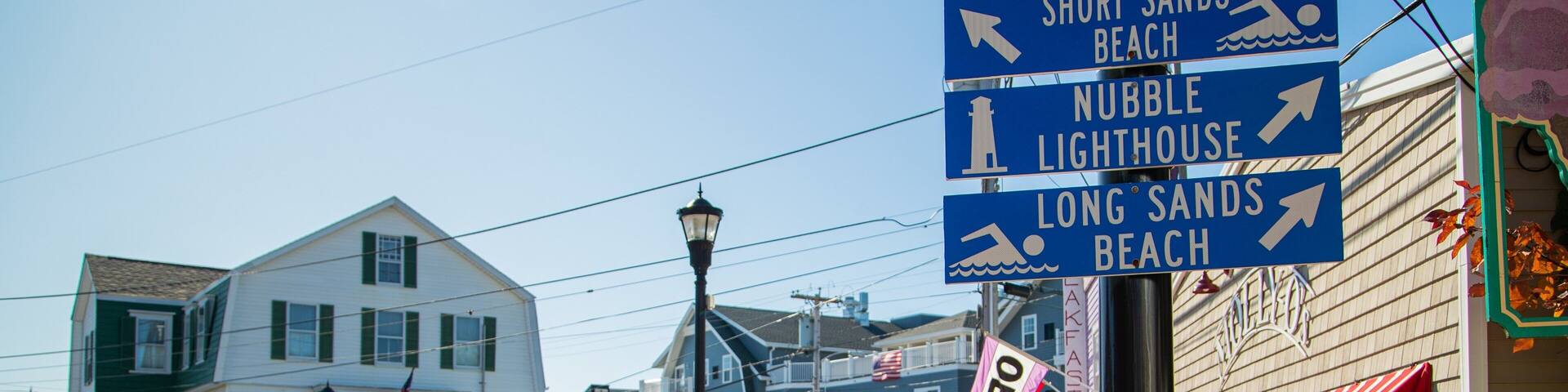 Une ville côtière où l’on aperçoit une Jeep garée dans la rue, un panneau indicateur indiquant la direction des plages et d’un phare, ainsi qu’un bâtiment sur lequel est affiché un panneau « OUVERT ».