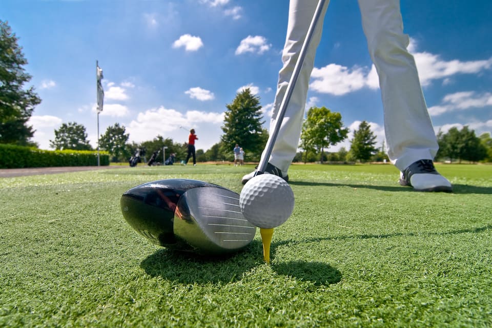 Una pelota de golf sobre el tee en un campo verde con árboles y una bandera al fondo.