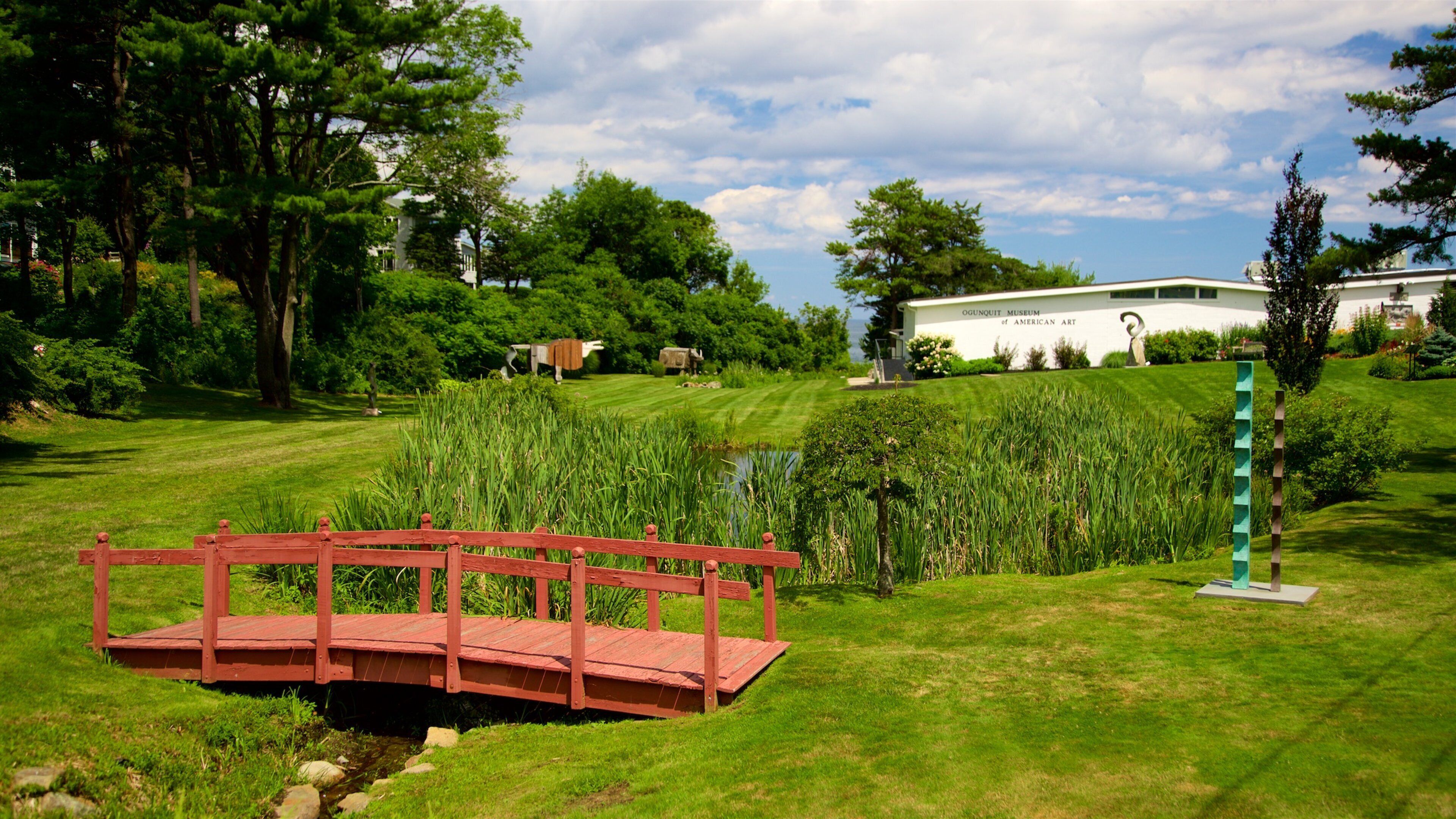 A red wooden bridge over a stream, surrounded by lush greenery and a white building labeled "Ogounquoit Museum of American Art" in the background.