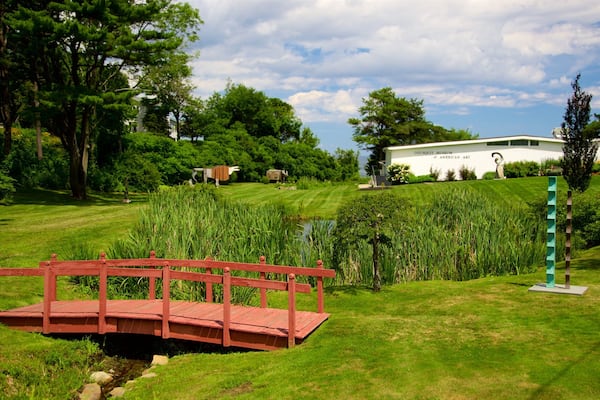 Un pont en bois rouge enjambant un ruisseau, entouré d’une végétation luxuriante, et, au fond, un bâtiment blanc portant l’inscription « Musée d’art américain Ogounquoit ».