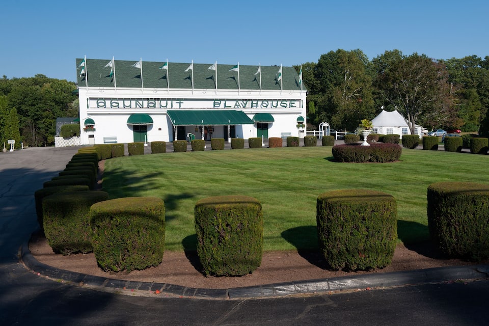 A building labeled "GUNNERT PLAYHOUSE" with a green roof, surrounded by a well-manicured lawn and hedges.