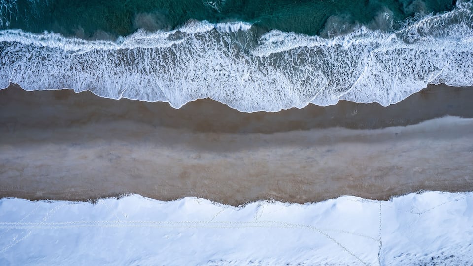 An aerial view of a beach with waves crashing onto the shore and snow on the sand.