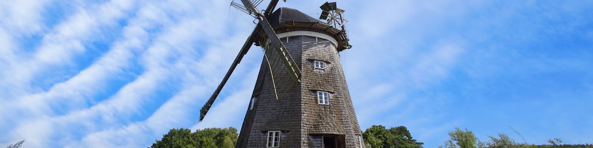 A traditional windmill with a wooden structure, set against a backdrop of a clear blue sky and lush greenery.