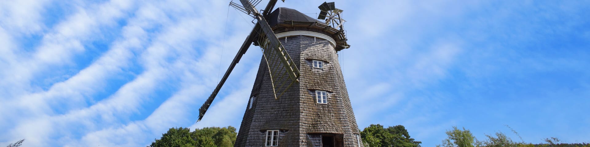 A traditional windmill with a wooden structure, set against a backdrop of a clear blue sky and lush greenery.