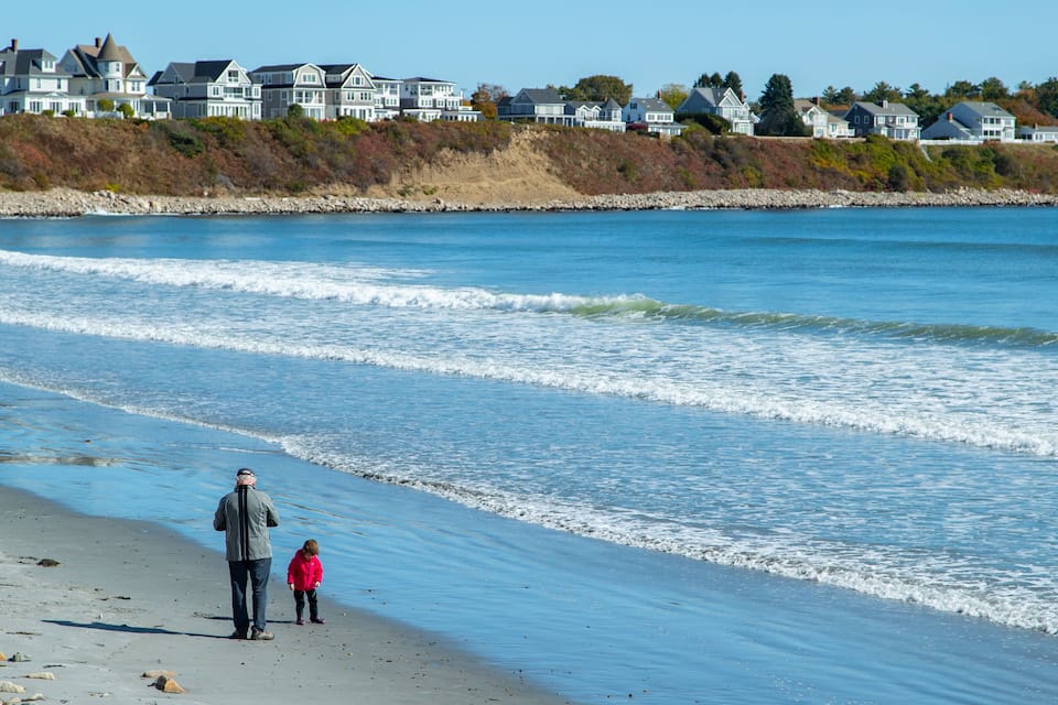 A beach with waves, a child in a pink jacket, and houses in the background.