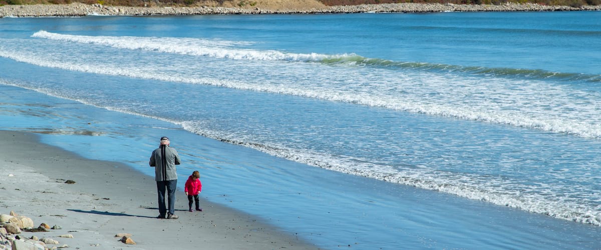 A beach with waves, a child in a pink jacket, and houses in the background.