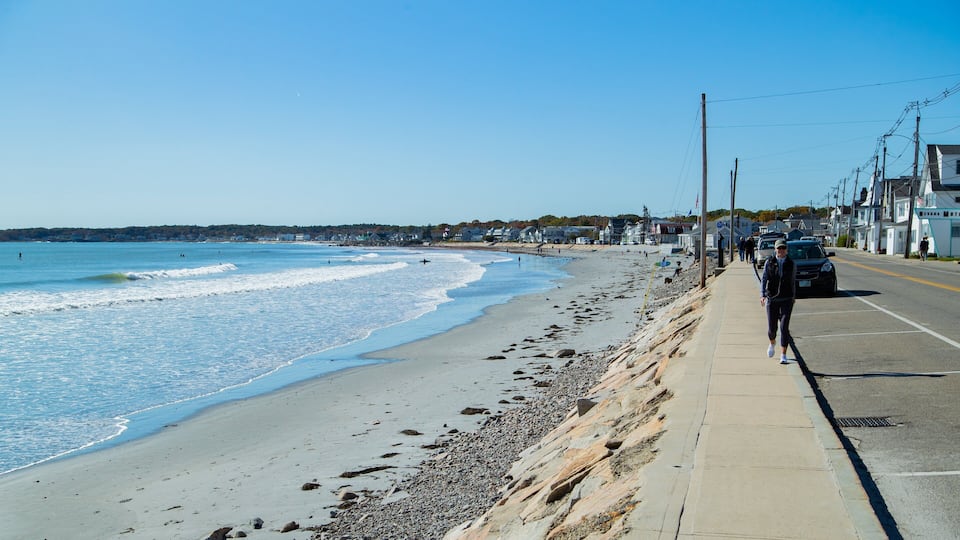 A beach with waves, a rocky shoreline, a sidewalk, and buildings in the background.