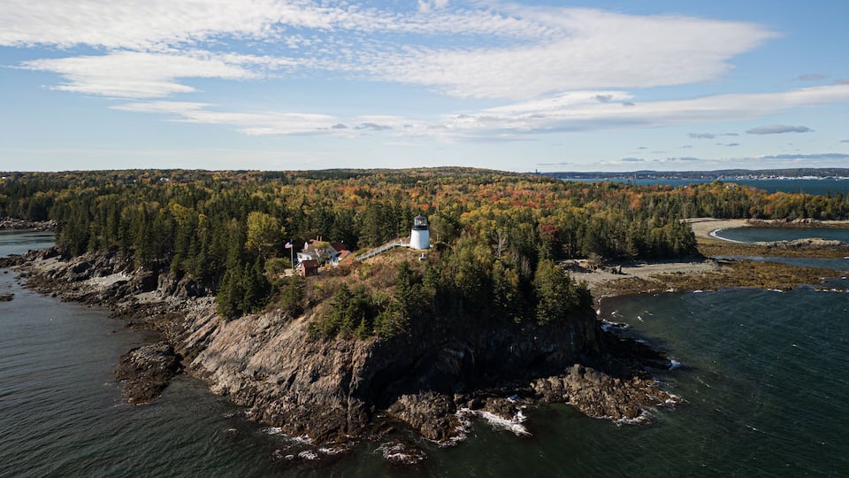 A lighthouse on a rocky island surrounded by autumn-colored trees and a calm sea.