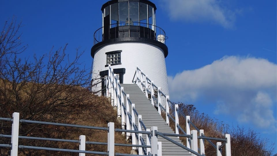 A lighthouse with a white exterior and black top, surrounded by a white fence and stairs leading up to it.