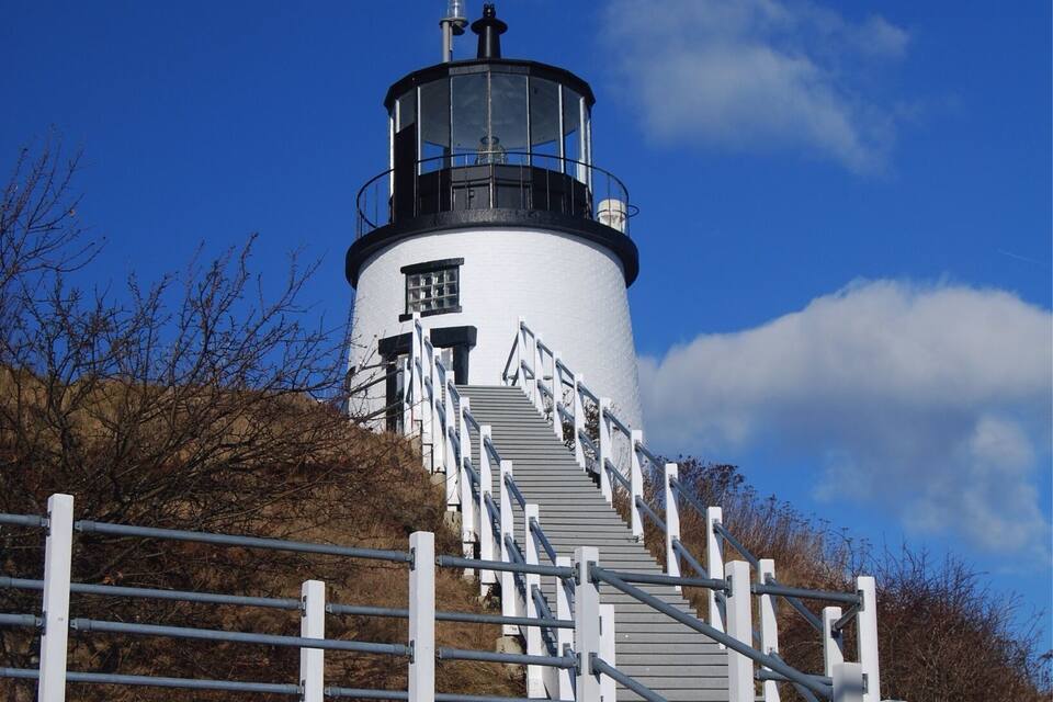 A lighthouse with a white exterior and black top, surrounded by a white fence and stairs leading up to it.