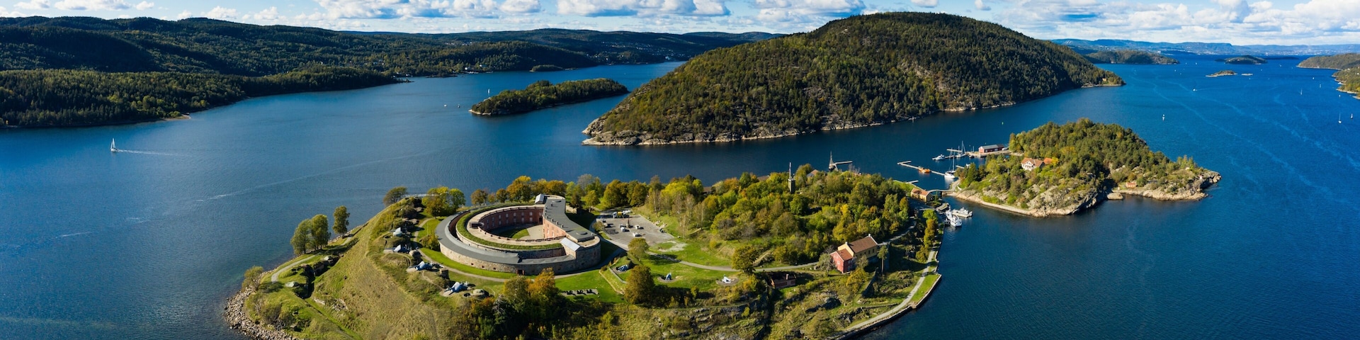 An aerial view of a large lake with several islands, one featuring a circular fortress-like structure surrounded by greenery.