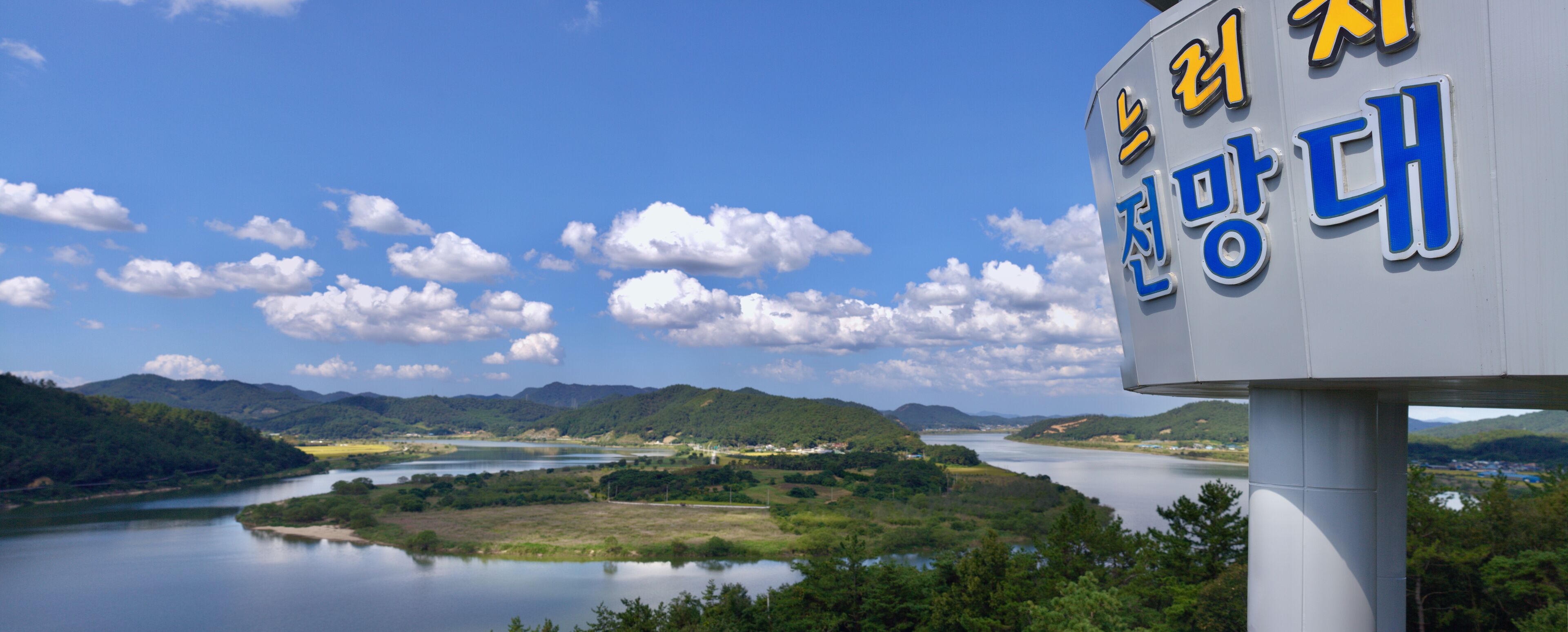 A sign with Korean text, a river, and a mountainous landscape.