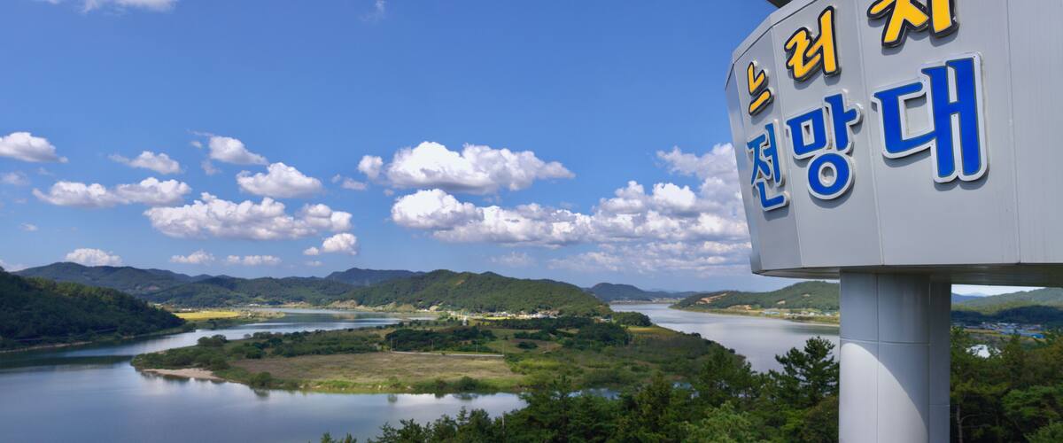 A sign with Korean text, a river, and a mountainous landscape.
