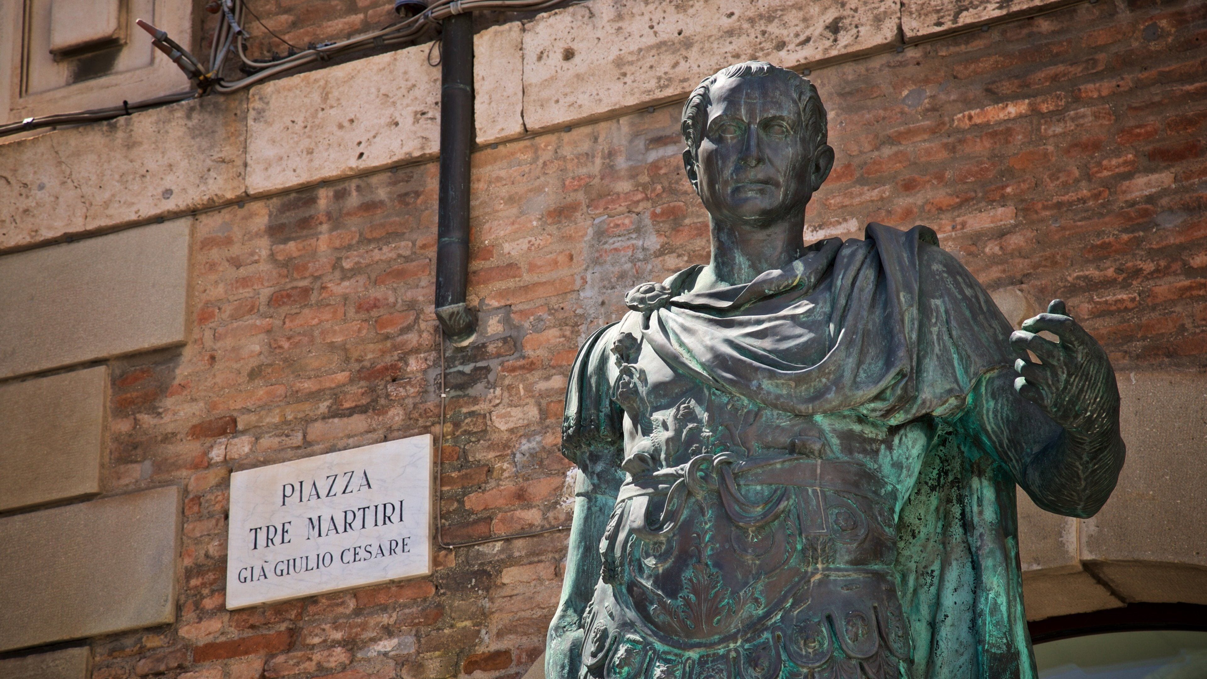 A bronze statue of a historical figure with a sign reading "Piazza Tre Martiri Gai Giulio Cesare".