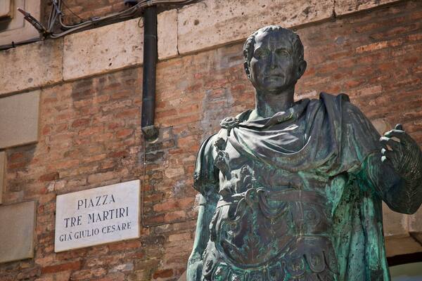 Une statue en bronze représentant une figure historique, avec une plaque indiquant : « Piazza Tre Martiri, Gaius Julius Caesar ».