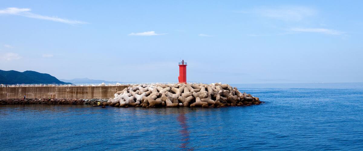 Un faro rojo sobre un espigón rocoso, con aguas azules tranquilas y un cielo despejado.