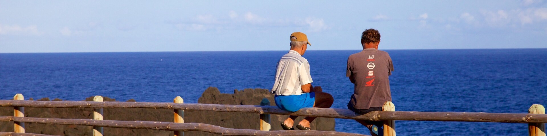 Dos personas sentadas en una valla de madera con vista al océano.