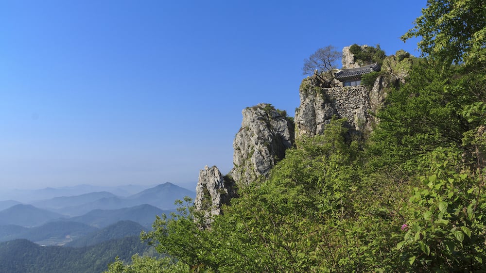 Ein traditionelles Gebäude thront auf einem steilen Felsen, umgeben von üppigem Grün und fernen Bergen.