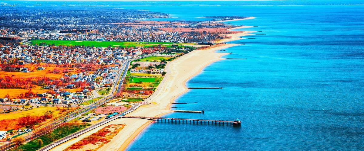 An aerial view of a coastal city with a long beach, a pier, and a highway running parallel to the shore.