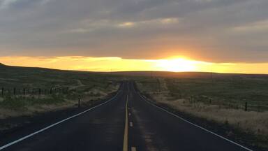 A straight road with a yellow center line, flanked by grassy fields and a cloudy sky.