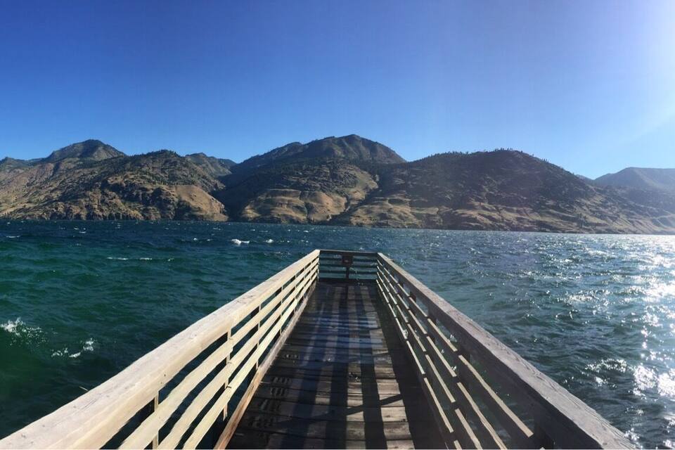Um píer de madeira se estende sobre um lago, com montanhas ao fundo.
