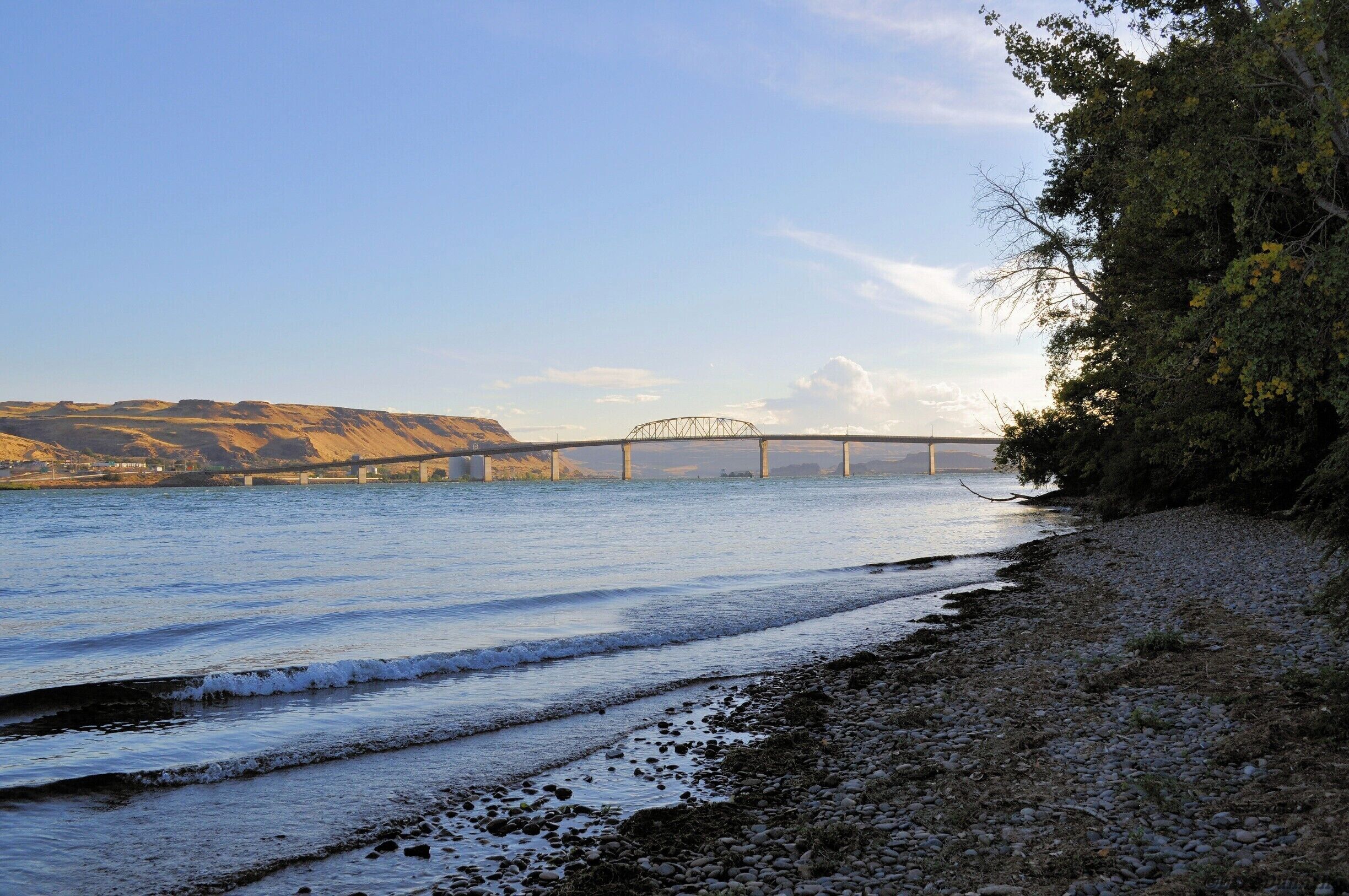 A river with a bridge, pebbly shore, and hills in the background.