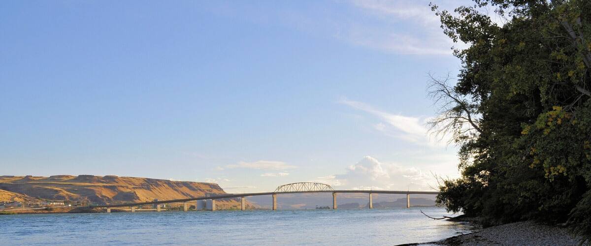 A river with a bridge, pebbly shore, and hills in the background.