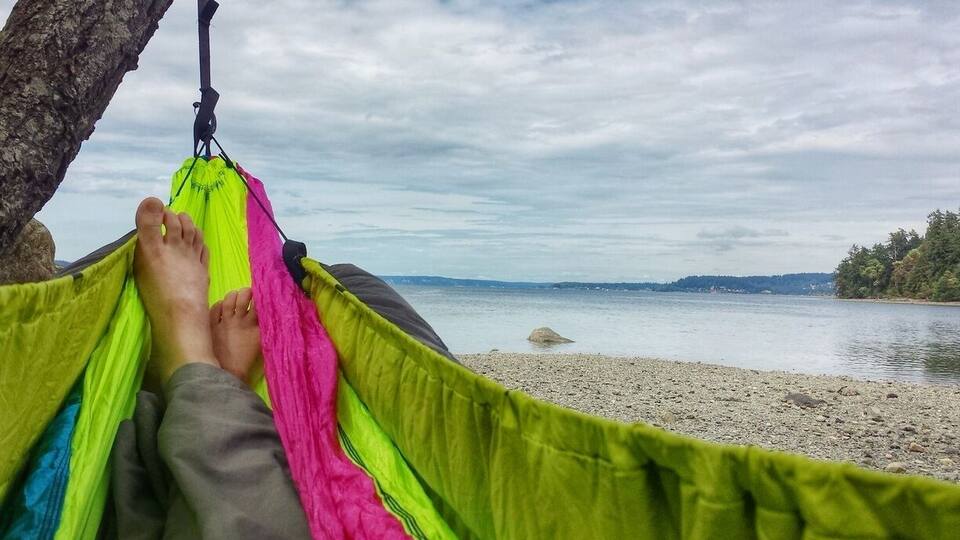 A hammock tied between trees on a pebbly beach with a lake and distant hills in the background.
