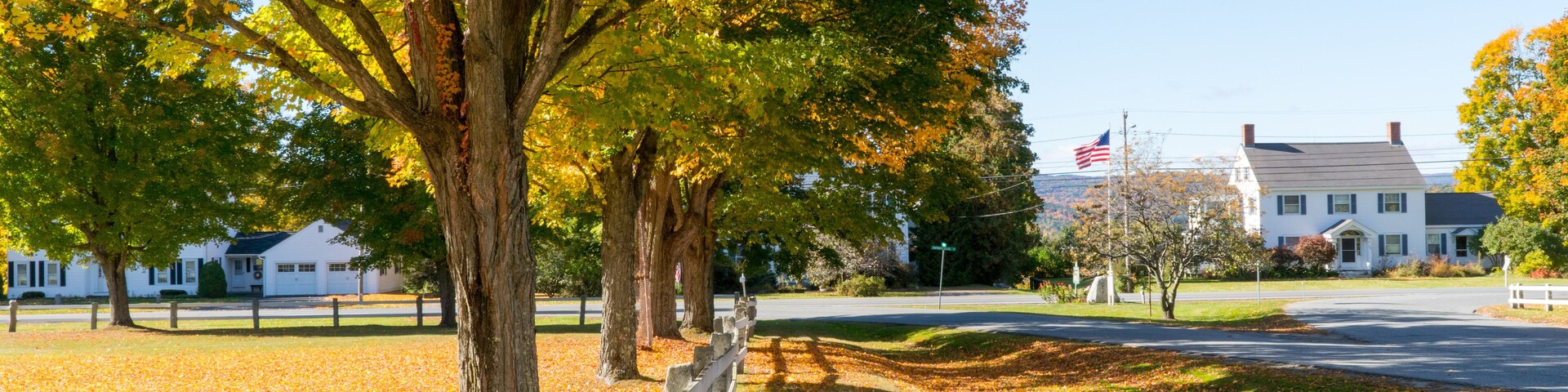 A park with autumn trees, a white fence, and a house with an American flag.