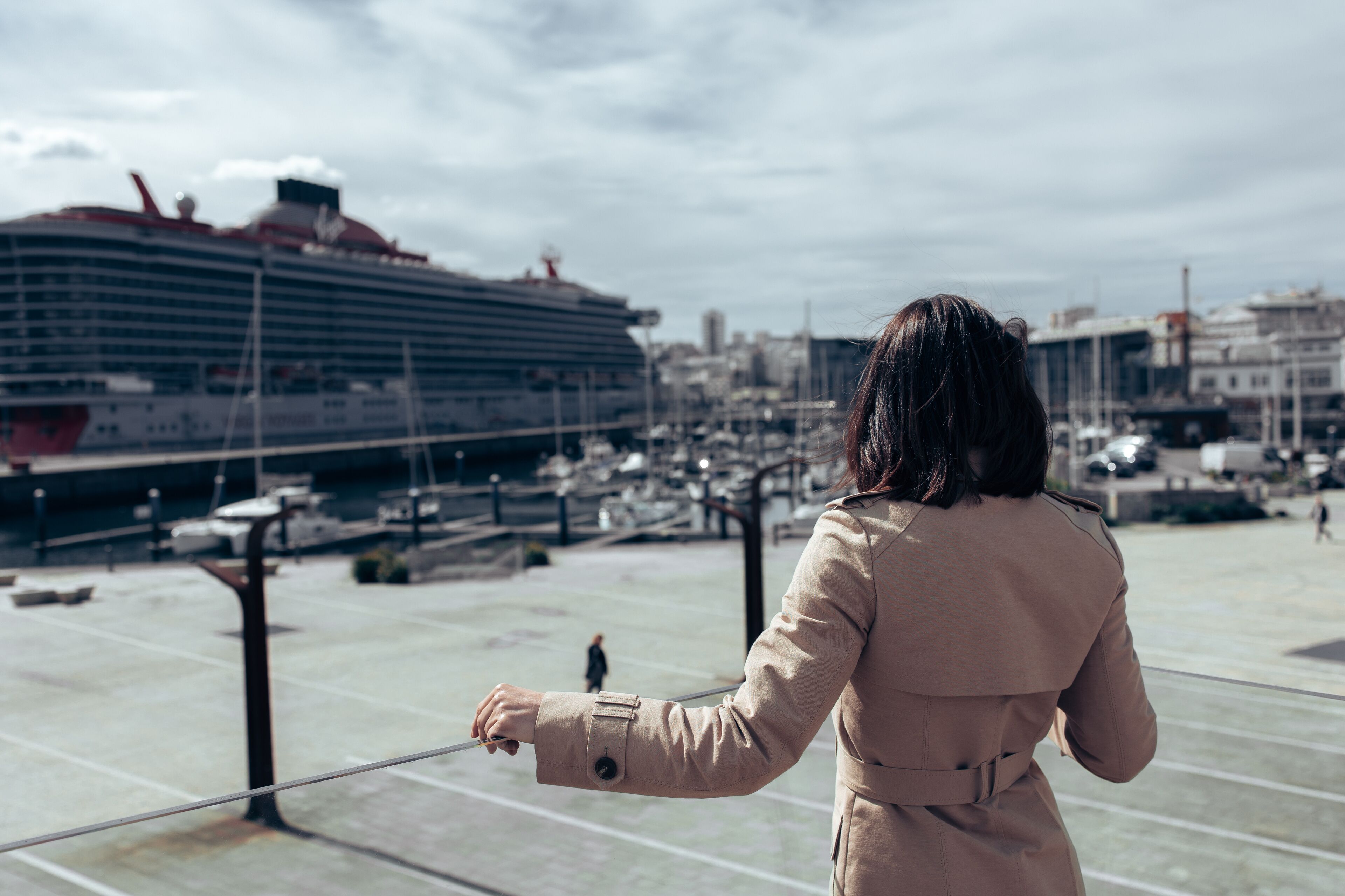 A girl, a brunette, in a beige cape, stands in the port and looks at the cruise ship. View from the back. The girl leans on a glass hedge. The sky is with clouds. There are many yachts in front of the
