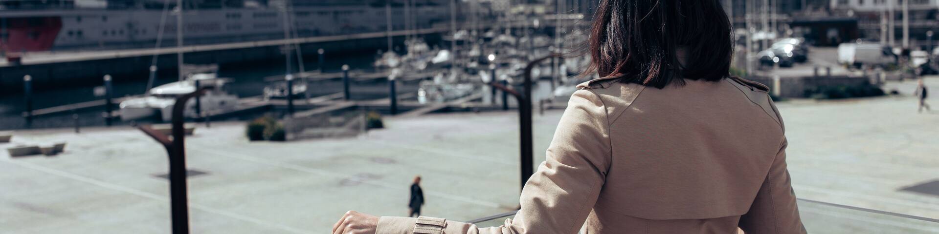 A girl, a brunette, in a beige cape, stands in the port and looks at the cruise ship. View from the back. The girl leans on a glass hedge. The sky is with clouds. There are many yachts in front of the