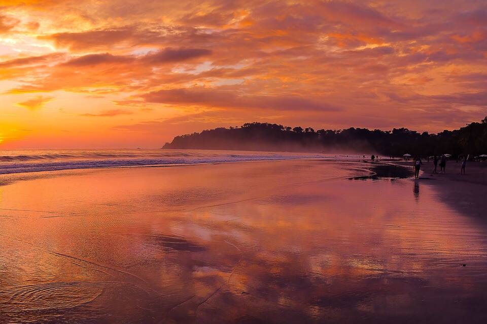 A beach at sunset with a calm sea, palm trees, and a distant island.
