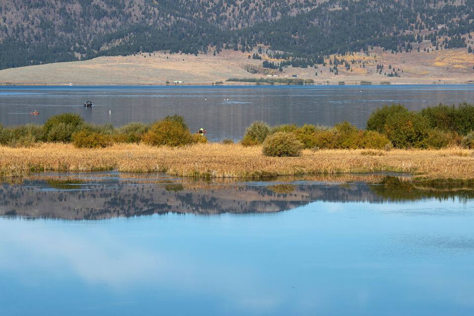 A calm lake with a clear reflection of the surrounding landscape, including distant hills and sparse vegetation.