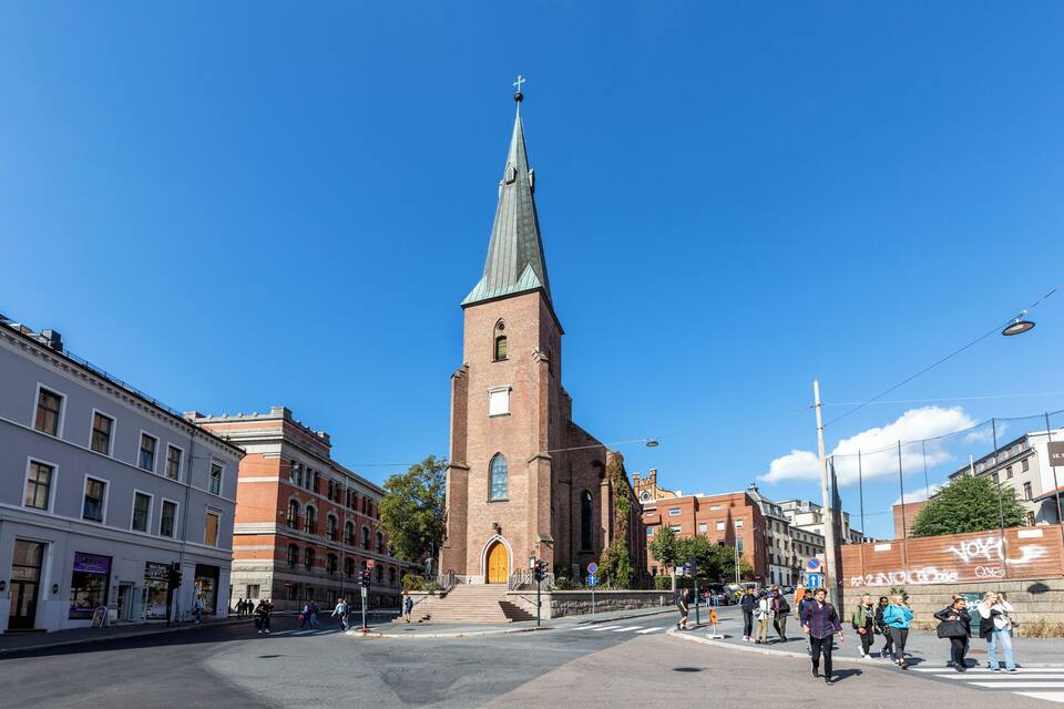 A church with a tall spire and a brick tower, surrounded by buildings and a clear blue sky.