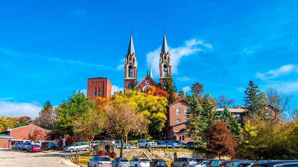 A church with two tall spires, surrounded by autumn trees and parked cars.