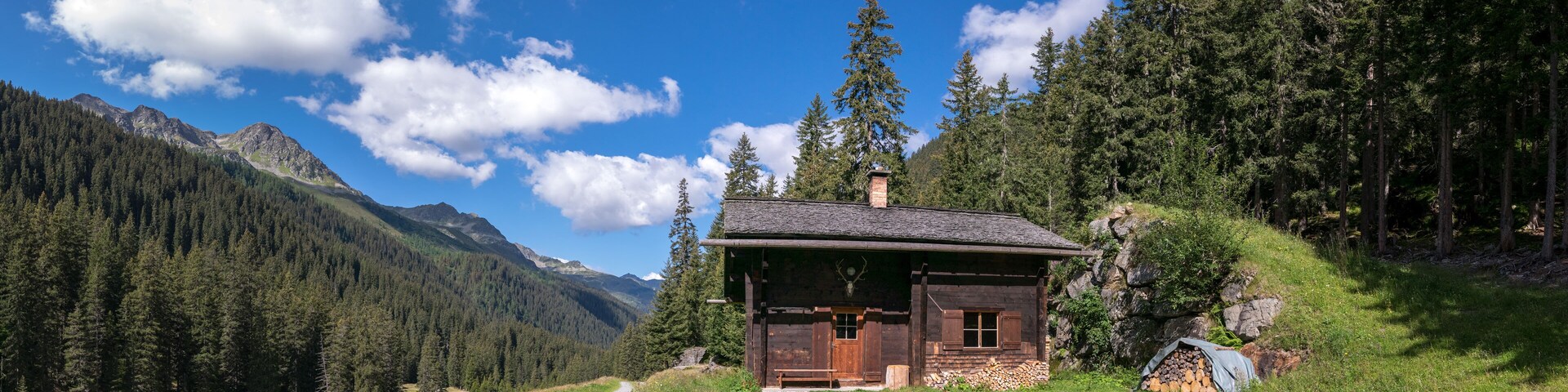 A wooden cabin in a forested valley with mountains in the background.