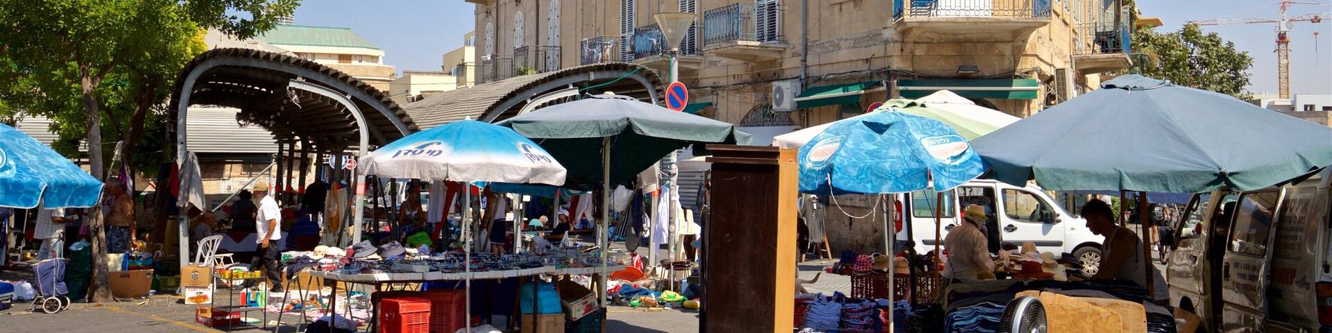 A bustling outdoor market with various stalls selling goods, surrounded by historic buildings and a clear blue sky.