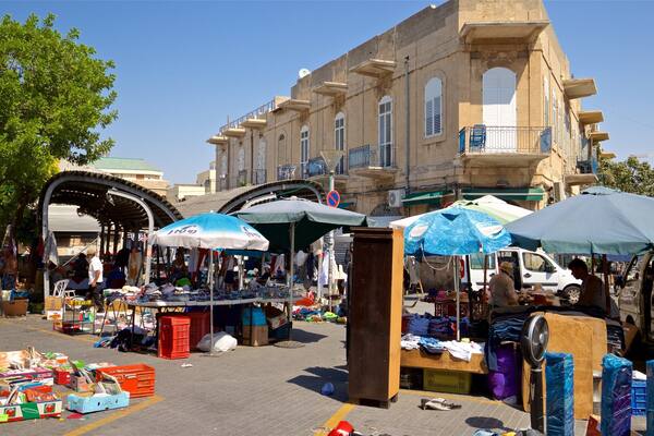 Um mercado ao ar livre movimentado, com diversos quiosques vendendo produtos, cercado por edifícios históricos e um céu azul.