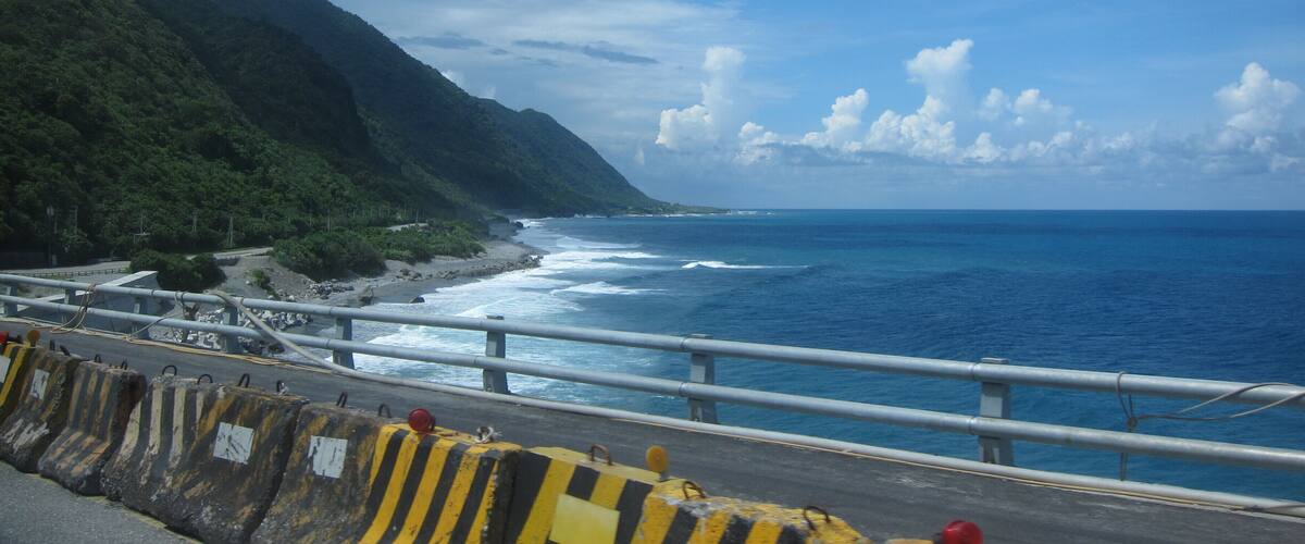 A coastal road with yellow and black barriers, a beach, and a mountainous landscape under a partly cloudy sky.