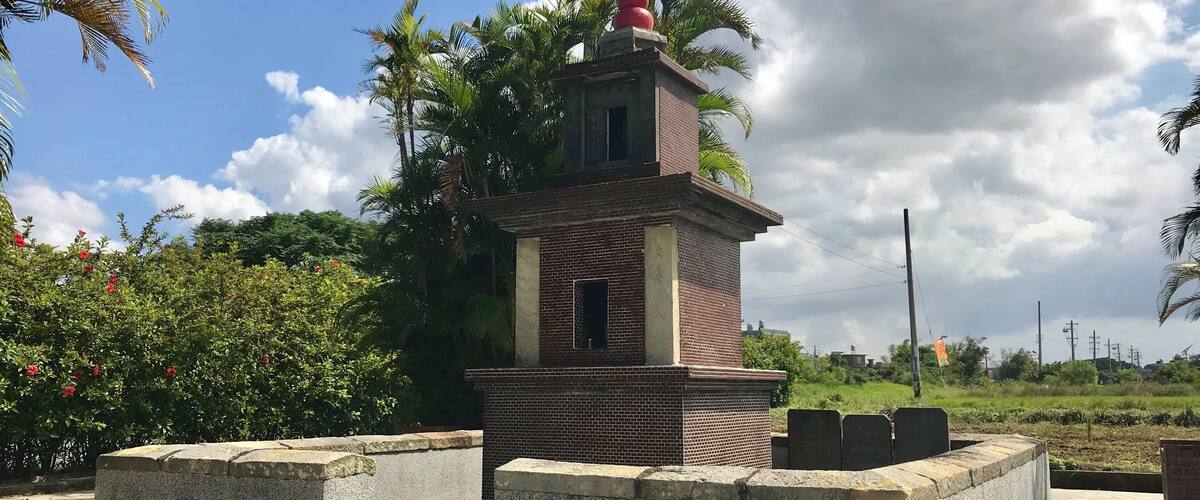A small brick monument with a statue on top, surrounded by greenery and a paved area.