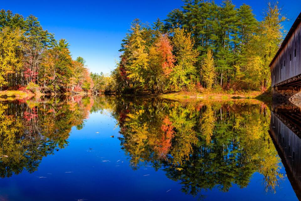 A wooden covered bridge over a calm river reflecting autumn trees.