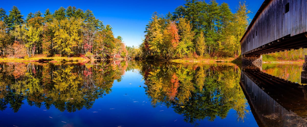 A wooden covered bridge over a calm river reflecting autumn trees.