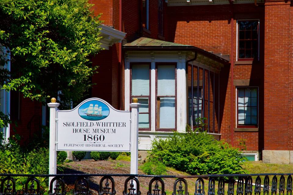A sign for the Skolfield-Whittier House Museum, dating back to 1860, with a brick building in the background.