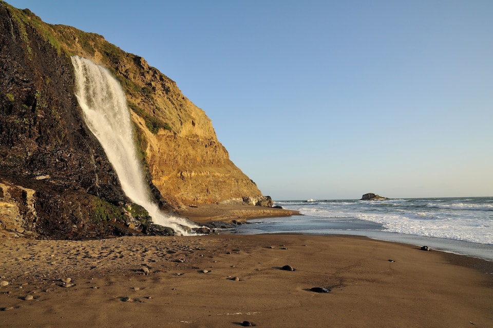 A coastal landscape with a waterfall cascading down a cliff, a sandy beach, and the ocean in the background.