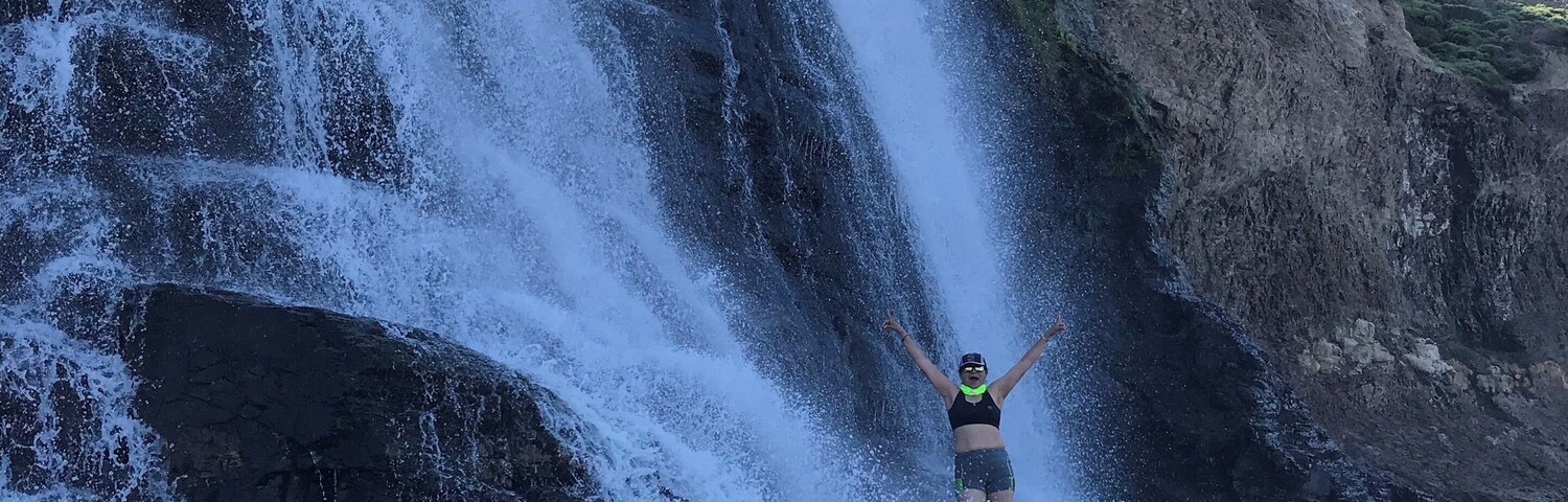 Eine Person steht vor einem Wasserfall, die Arme erhoben.