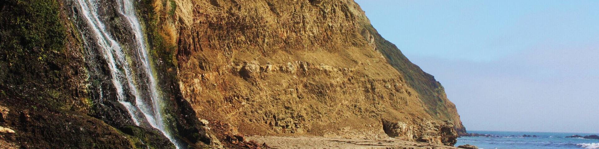 A coastal cliff with a waterfall and a sandy beach.