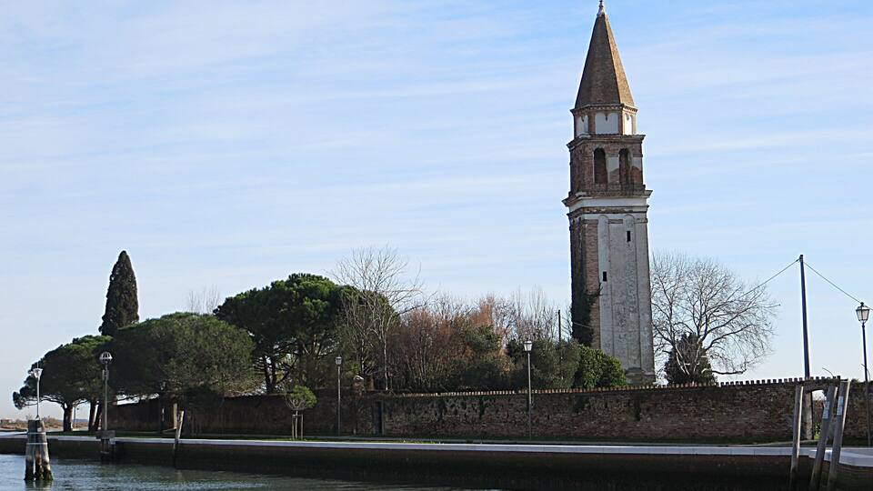 A historic tower by a calm river, surrounded by trees and a stone wall.
