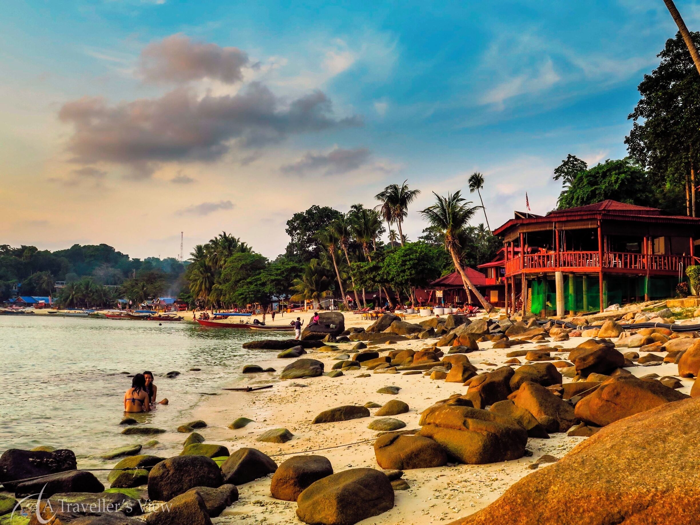 A beach with large rocks, palm trees, and a red building.